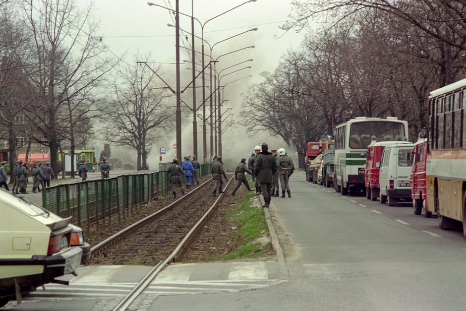 Ulica, na poboczu sznur samochodów, w środku tory tramwajowe, na nich strażacy. W tle kłęby dymu