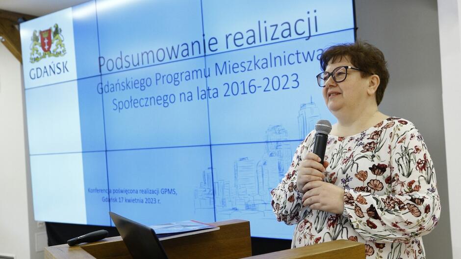 Kobieta w okularach przemawiająca do mikrofonu z planszą z napisem podsumowanie Gdańskiego Programu Mieszkalnictwa Społecznego 2016-2023