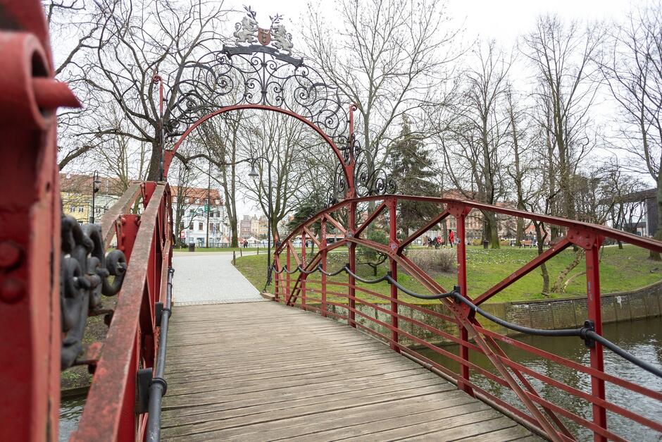 stalowy most w kolorze czerwonym. Na pierwszym planie powierzchnia mostu wykonana z desek, w tle widoczny park