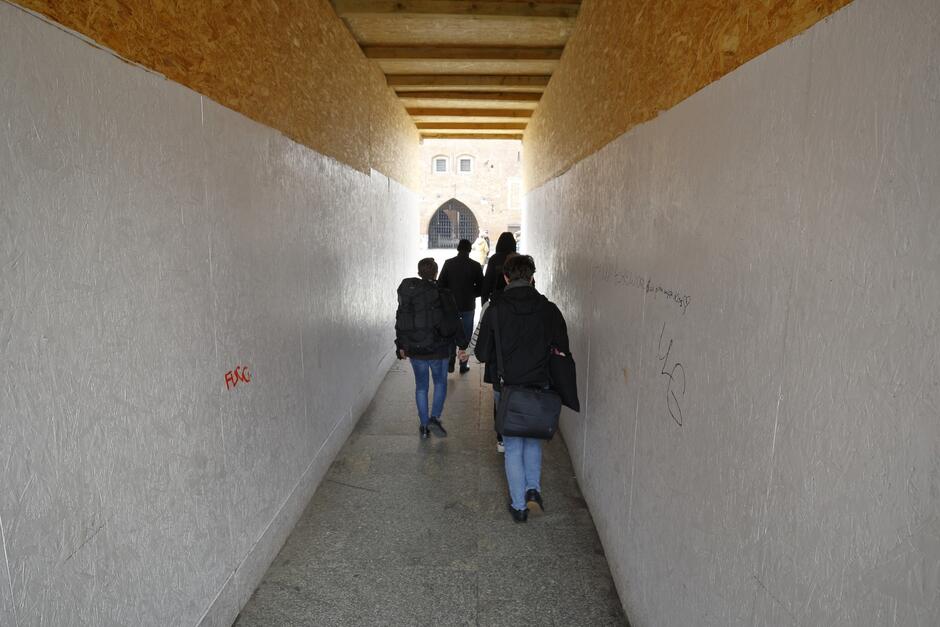 Ludzie przechodzą przez długi korytarz o prostokątnym przekroju - ściany i sufit zrobione z płyt pilśniowych