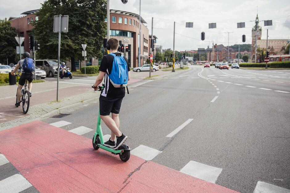 na zdjęciu chłopak w letnim stroju jadący na hulajnodze po ścieżce rowerowej, przejeżdża przez ulicę