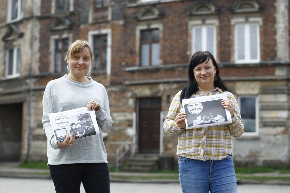 dwie kobiety trzymają w rękach gazety, za nimi kamienica