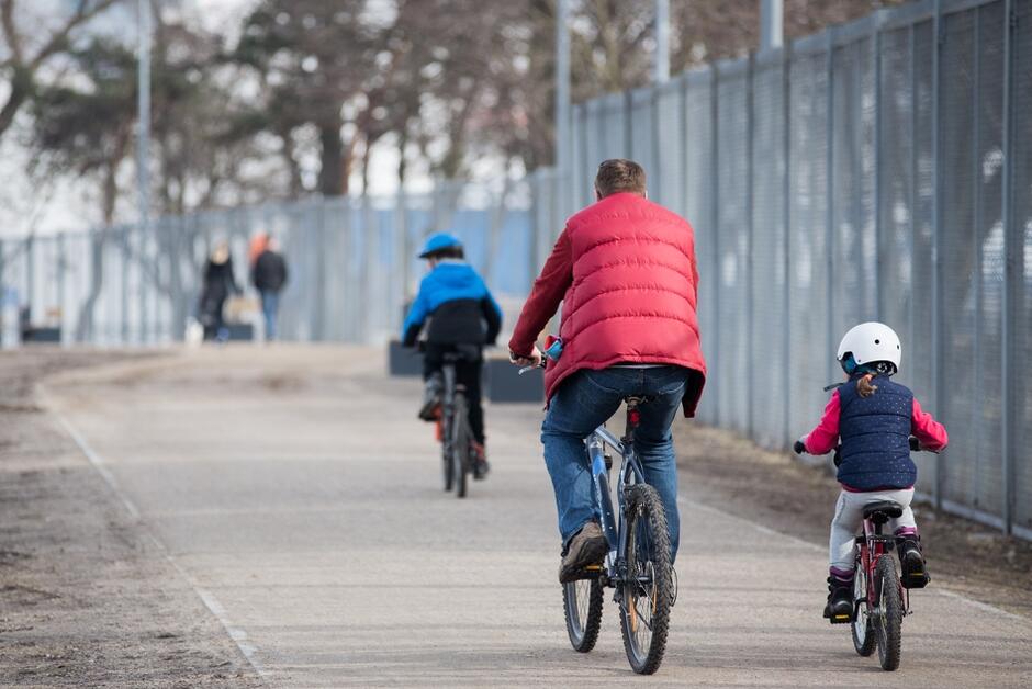 na zdjęciu dwoje dzieci i jeden dorosły mężczyzna jadą na rowerach, mają wiosenne kurtki na sobie, wszyscy jadący są plecami do fotografującego