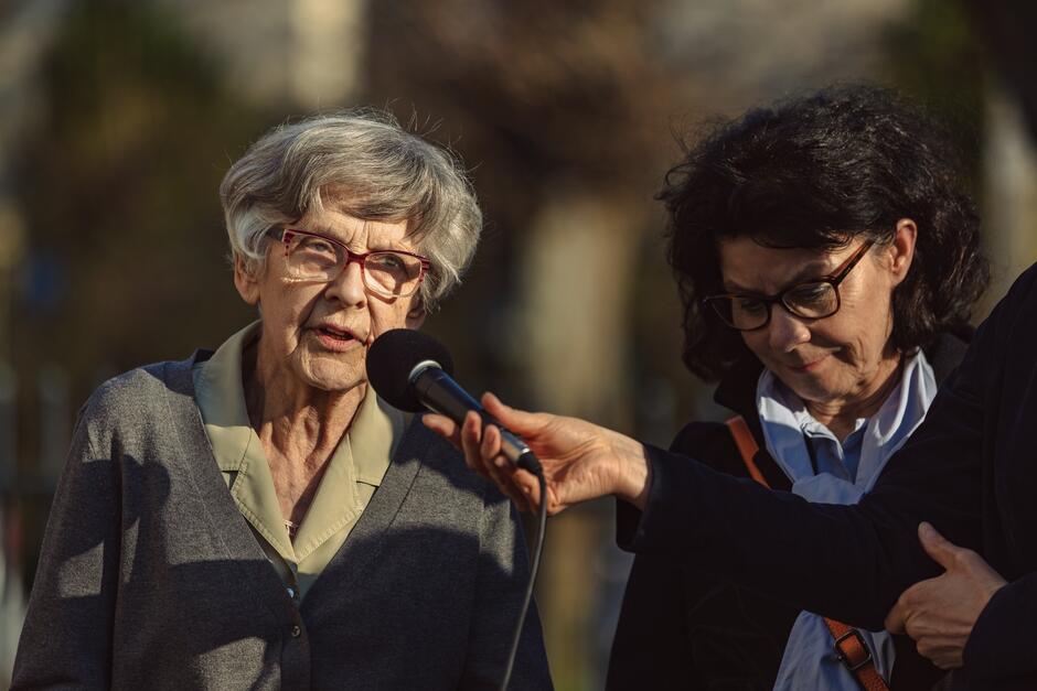na zdjęciu dwie starsze kobiety, jedna w szarej bluzce mówi do mikrofonu, druga stoi obok niej