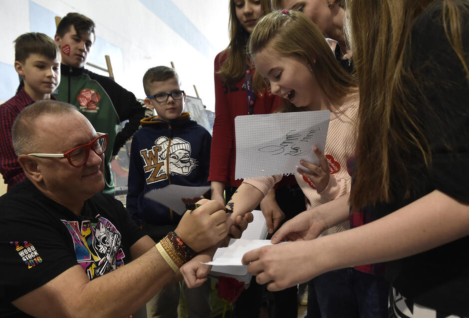 Mężczyzna w okularach rozdający dzieciom autografy, jednej dziewczynce składa podpis flamastrem na ręce