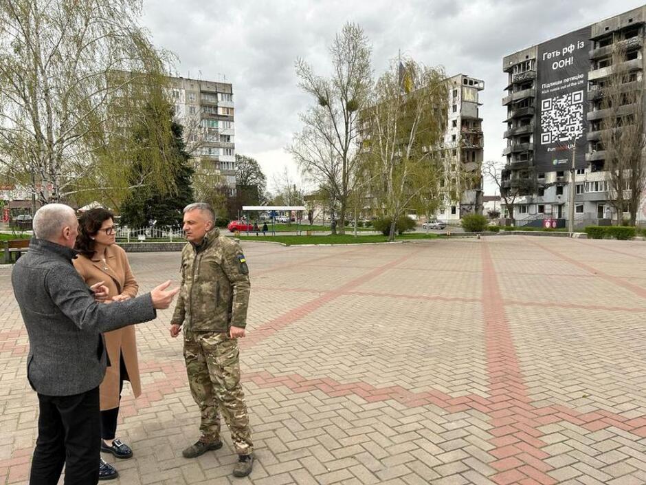 kobieta w średnim wieku stoi na placu z i rozmawia z dwoma mężczyznami w tle zniszczone przez pociski bloki