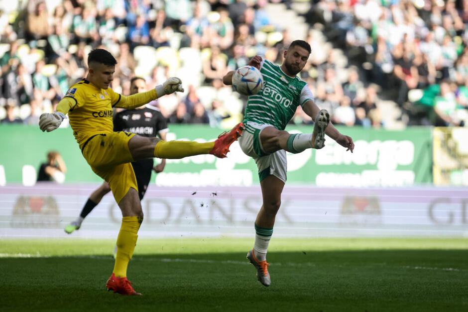 Bramkarz w żółtym stroju, piłkarz w biało-zielonej koszulce. Piłka w powietrzu, obaj mają wyciągniętą do góry nogę