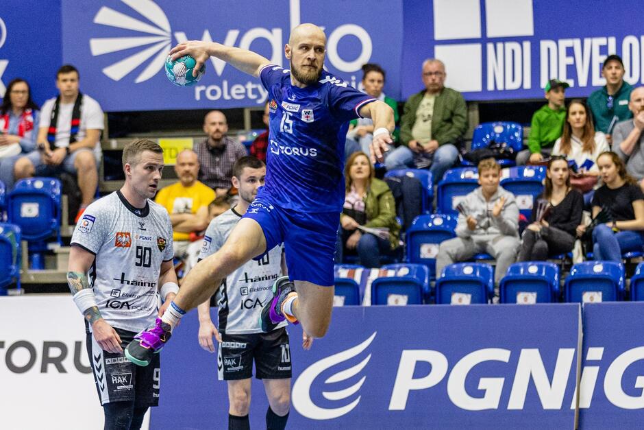 Łysy mężczyna w niebieskim stroju w wyskoku z piłką. Za nim dwóch zawodników w białych koszulkach, w tle trybuny z kibicami