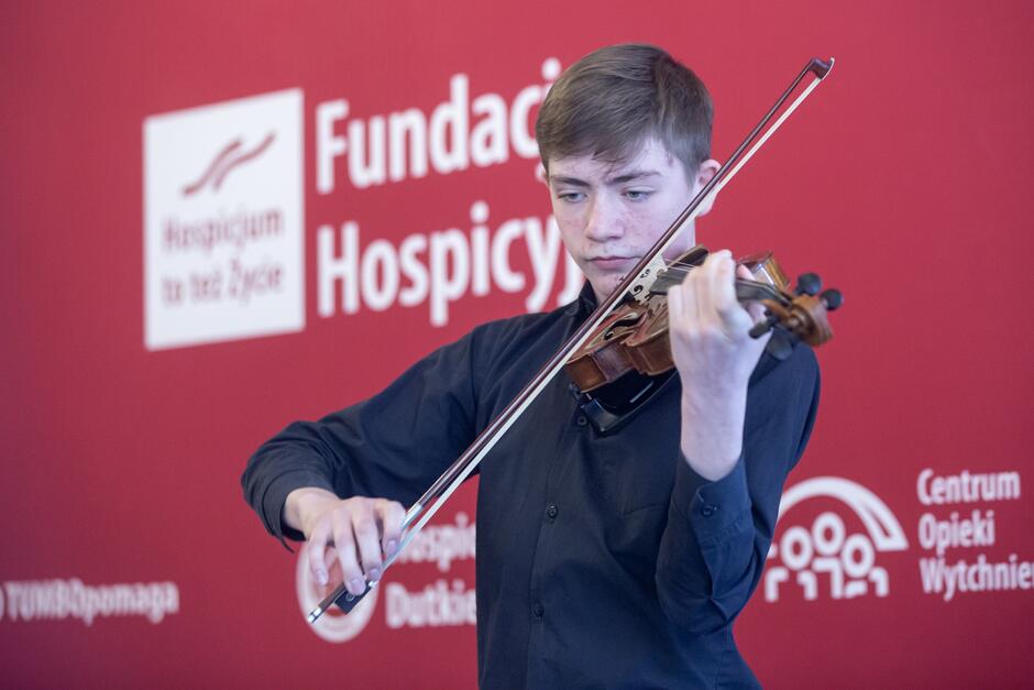 Chłopiec w wieku około lat 15-16, grający na skrzypcach. Ubrany w czarną koszulę, jest szatynem 