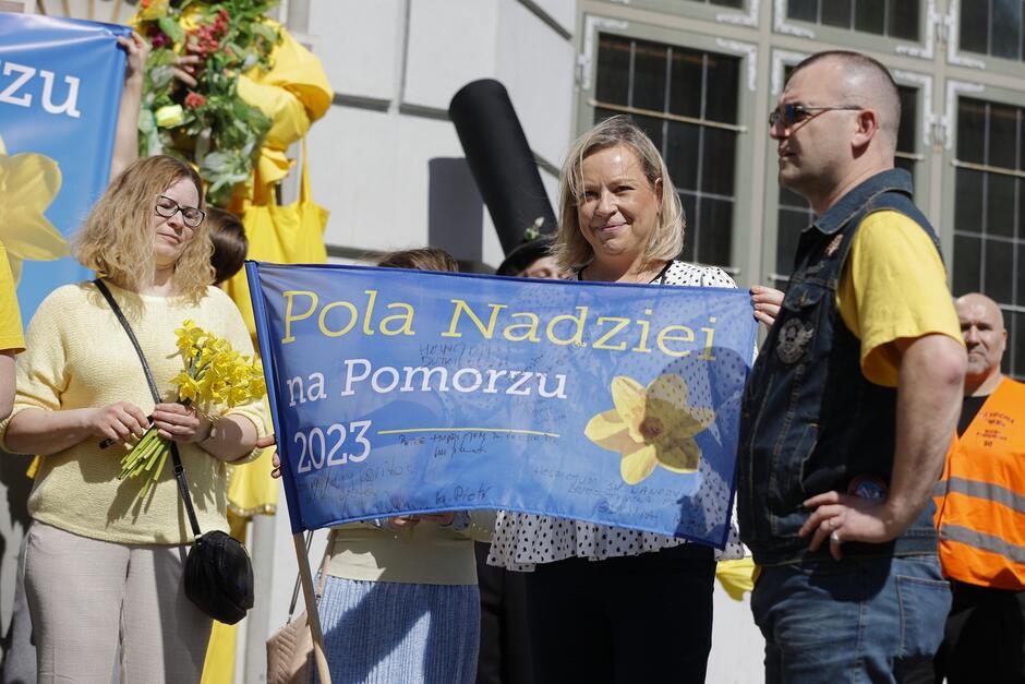 na zdjęciu blondynka w średnim wieku trzyma w rękach niebieską flagę, uśmiecha się do fotografującego, obok niej stoi kobieta i mężczyzna