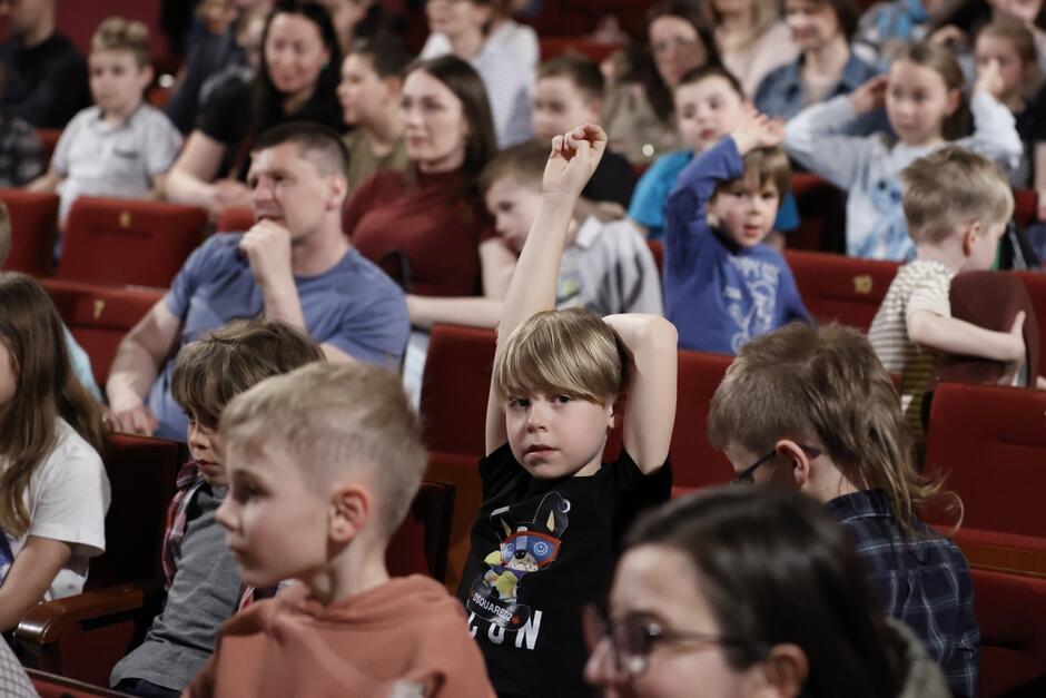 Małe dzieci siedzą na czerwonych teatralnych fotelach. Dwoje z nich podnosi rękę