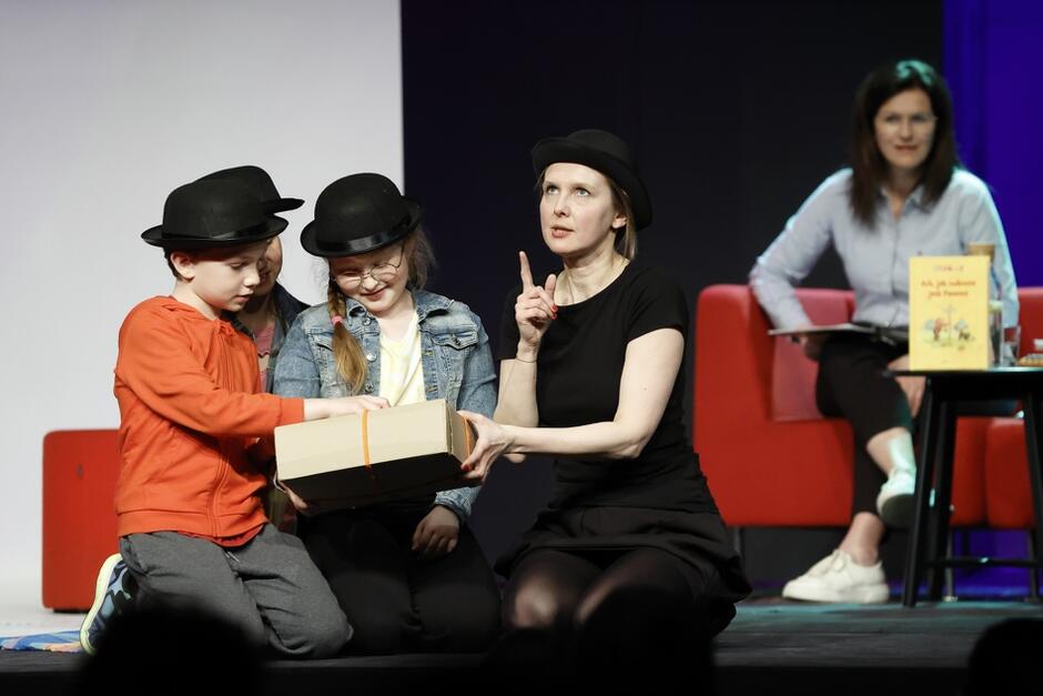 Troje dzieci siedzi na scenie. Mają na głowie czarne meloniki i próbują razem otworzyć paczkę. W tle młoda kobieta siedzi na kanapie z mikrofonem i książką
