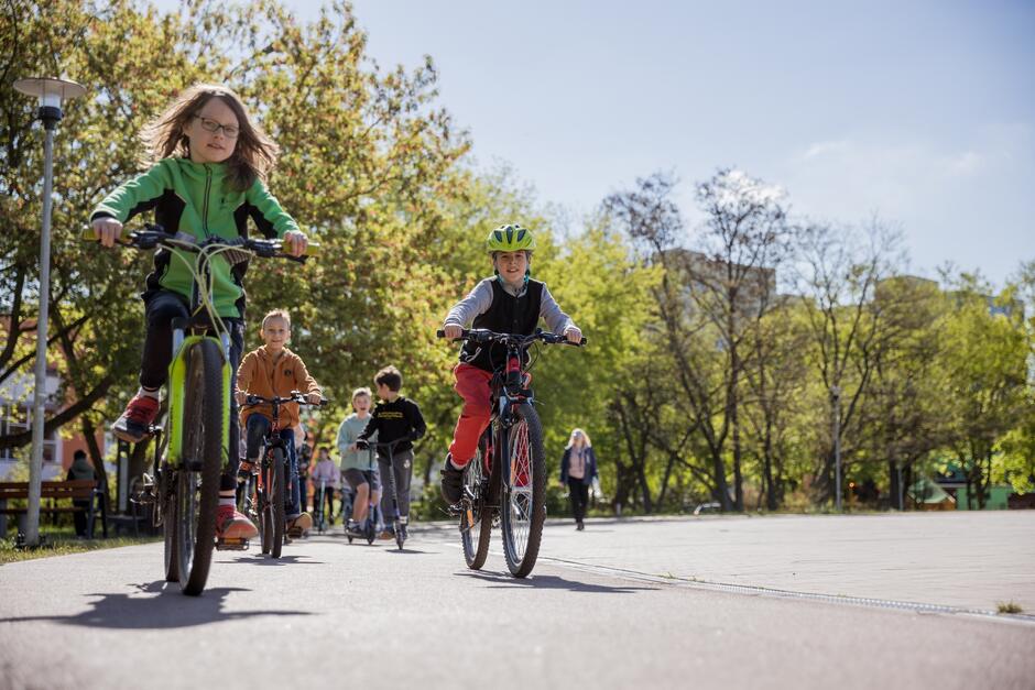 Dzieci na ścieżce rowerowej na rowerach i hulajnogach, po bokach zieleń