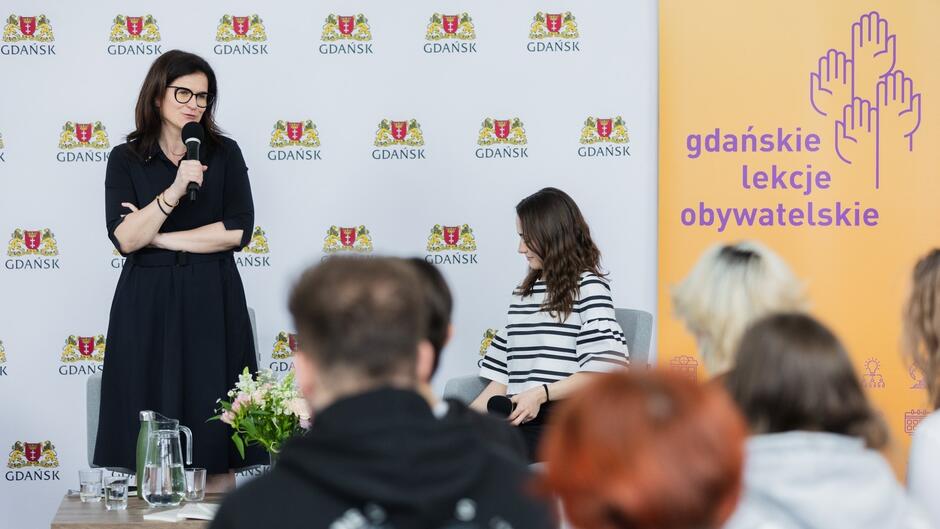 Kobieta w okularach przemawiająca do uczniów na tle planszy z napisem Gdańskie Lekcje Obywatelskie