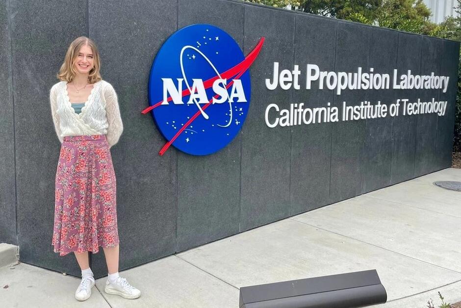Ładna młoda, szczupła dziewczyna o długich włosach stoi po prawej pozując do zdjęcia przy niebiesko-białym logo NASA, które jest po prawej. Widzimy całą jej postać. Dziewczyna jest w długiej i lekkiej spódnicy w drobne kwiatki, wpadającej w czerwień i w jasnym sweterku. Na nogach ma białe adidasy 
