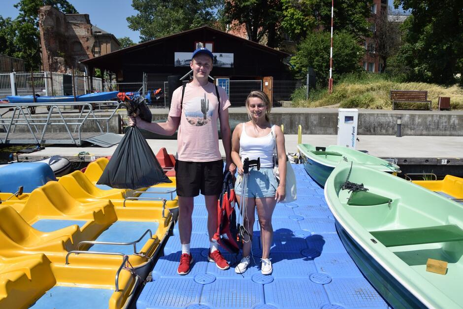 na zdjęciu młoda para, chłopak i dziewczyna, trzymają czarny worek na śmieci, i chwytaki, stoją na plastikowej platformie umieszczonej na wodzie, wokół małe łódki i kajaki