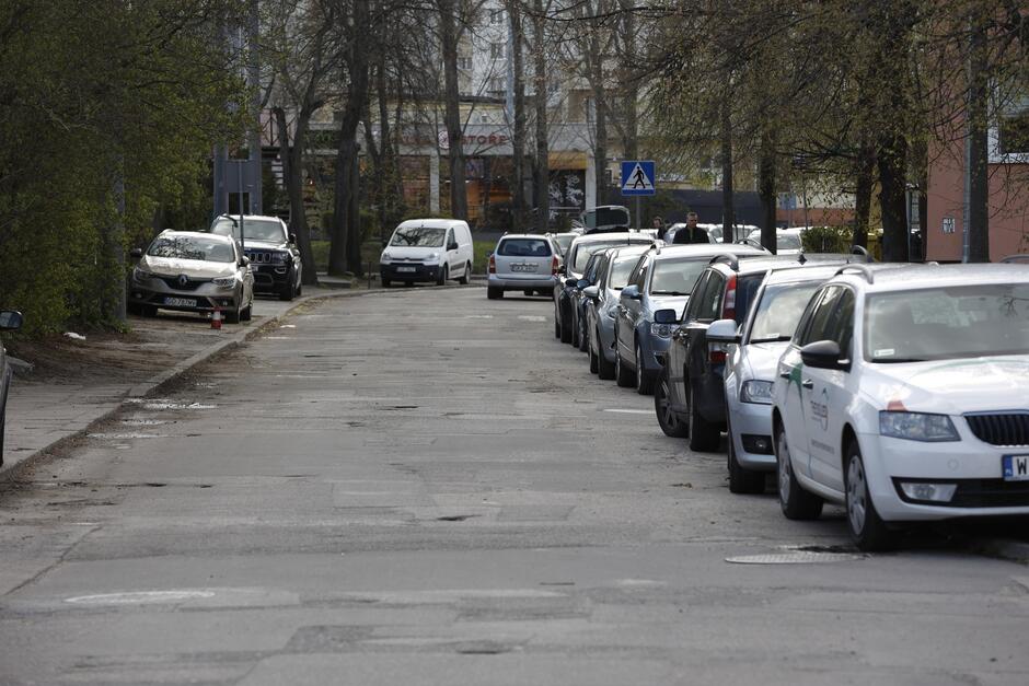 na zdjęciu fragment ulicy i sznur kilkunastu samochodów zaparkowanych jeden za drugim częściowo na chodniku