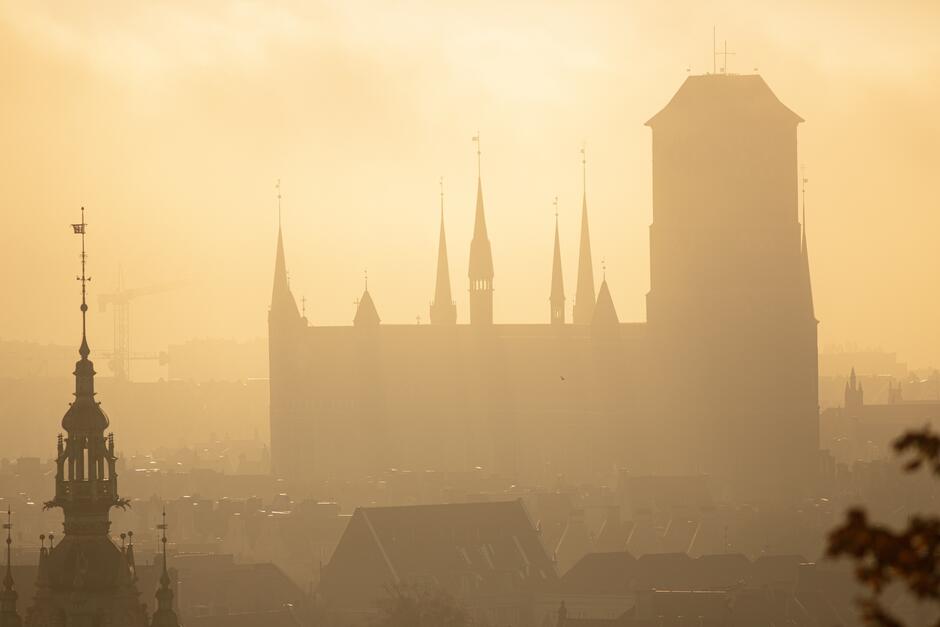 Sylwetka bazyliki Mariackiej, zdjęcie wykonane z góry Gradowej. Panuje mgła, przez którą próbuje przebić się światło słoneczne, co daje efekt złotej poświaty 