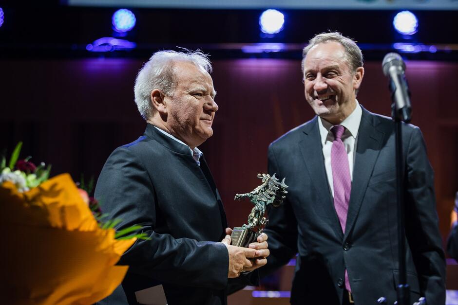 Dwa eleganccy mężczyźni w garniturach stoją na scenie przed mikrofonem, starszy z siwymi włosami trzyma statuetkę pomorskiego orła. Uśmiechają się 
