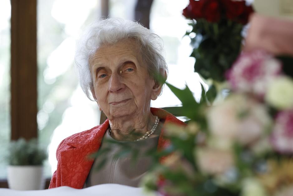 Zdjęcie przedstawia sędziwą panią w czerwonej bluzce, która siedzi przy stole zastawionym bukietami kwiatów