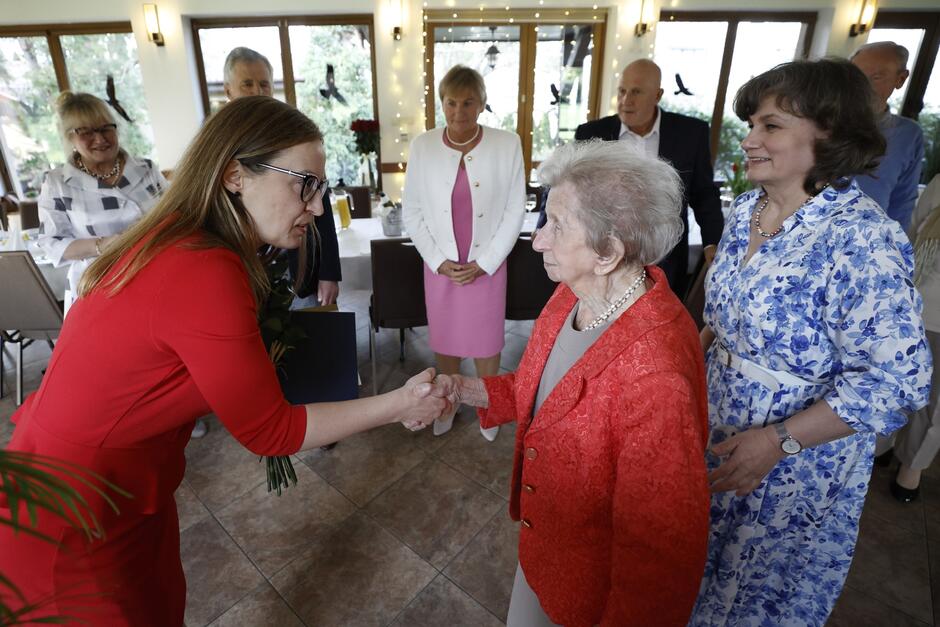 Dwie kobiety podają sobie rękę na powitanie. Po lewej wyższa, w średnim wieku, ubrana w czerwoną sukienkę. Po prawej - sędziwa, o siwych włosach, ubrana w popielatą bluzkę i czerwony żakiet   