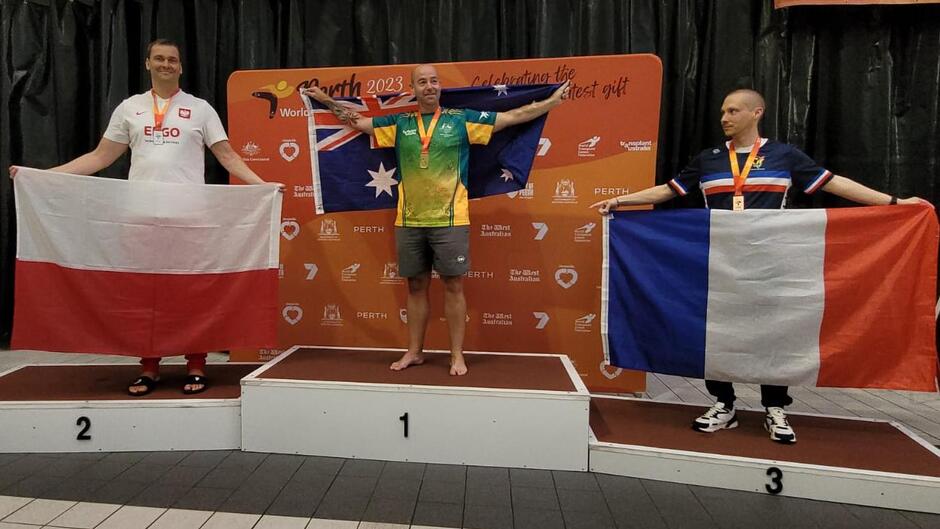 Trzech mężczyzn stoi na podium, w dłoniach nad głowami trzymają flagi narodowe 