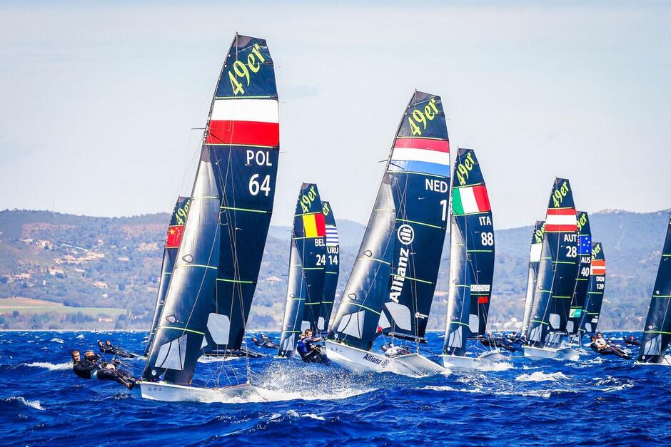 Jachty z wysokimi żaglami płyną obok siebie. Na czele łódka z żaglem z biało-czerwoną flagą