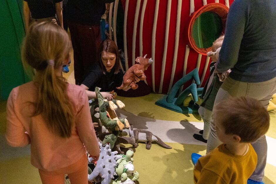 młoda kobieta siedzi przed stosem różnych pluszowych dinozaurów, wokół niej kilkoro dzieci w wieku od 3 do 6 lat