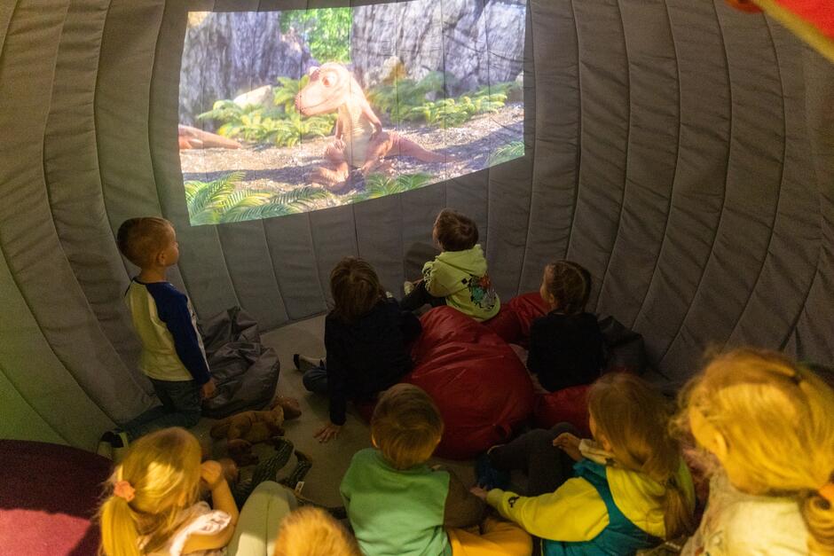 dzieci patrzą na ścianę, na której w małym ekranie widać model dinozaura
