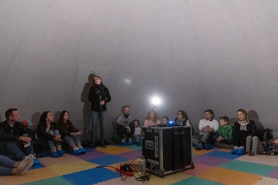 rodzice z dziećmi siedzą na podłodze, wokół ścian planetarium. Młoda kobieta, pracowniczka Hevelianum coś opowiada