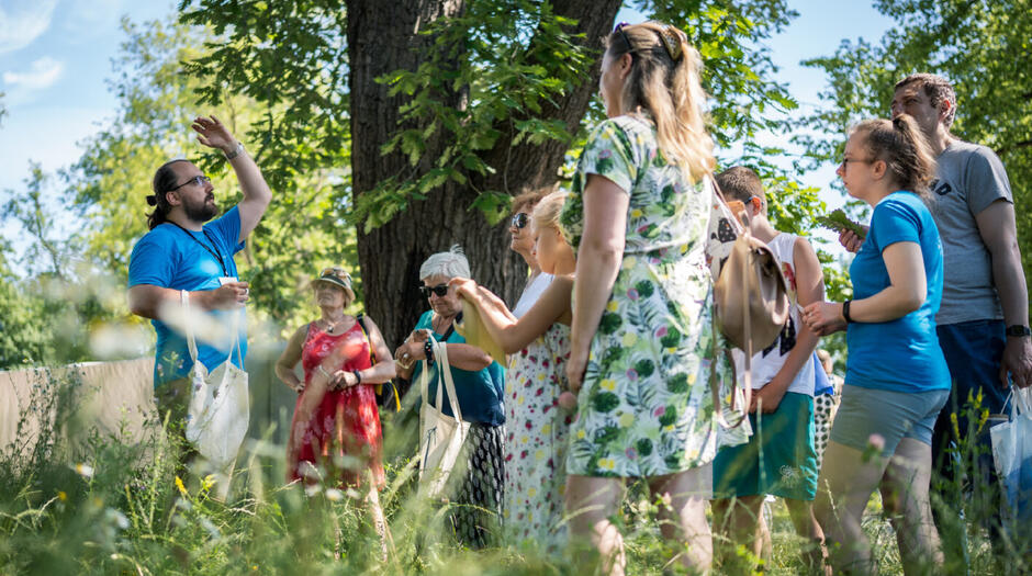 grupa ludzi w różnym wieku stoi na łące pod drzewem, młody mężczyzna wznosi rękę nad głowę