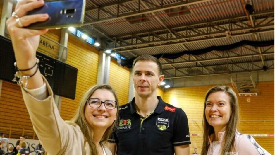 Mężczyzna w stroju sportowym, po jego bokach dwie uśmiechnięte kobiety, jedna z nich trzyma przed sobą telefon komórkowy, robiąc selfie