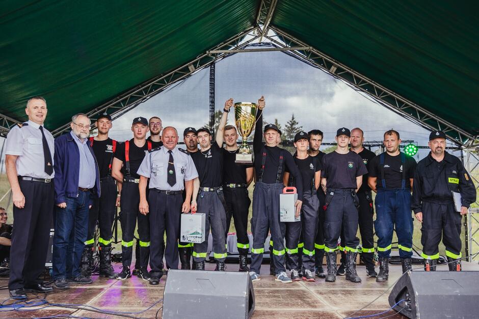 Grupa strażaków w ciemnych mundurach z pucharem za zwycięstwo w zawodach. Stoją pod zadaszeniem, który daje namiot w ciemnozielonym kolorze 