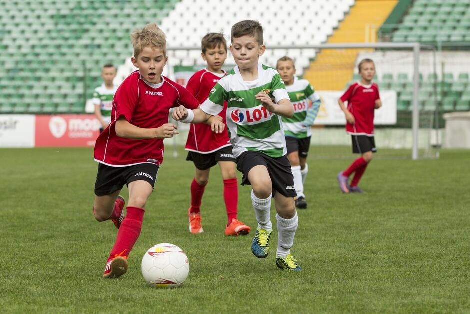 Chłopcy na murawie biegną za piłką. Na pierwszym planie w czerwonej koszulce, za nim w biało-zielonej