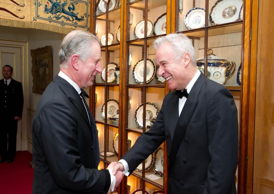Dwaj eleganccy i uśmiechnięci mężczyźni witają się uściskiem ręki w pałacowej sali. Po lewej jest książę Karol, po prawej - prof. Limon 