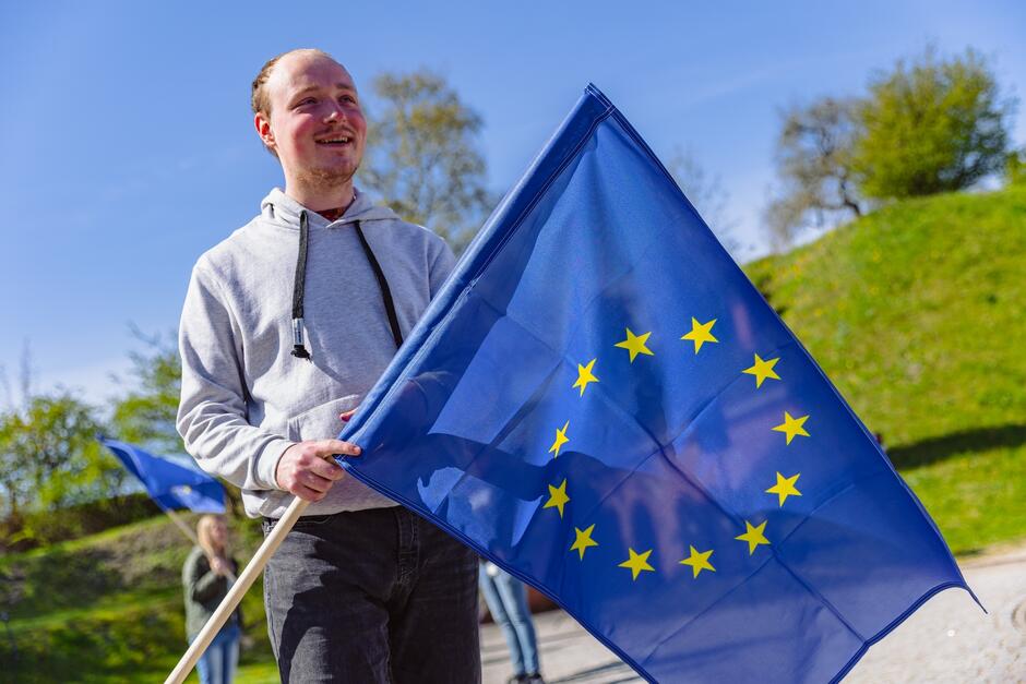 Młody uśmiechnięty mężczyzna trzyma w dłoniach flagę Unii Europejskiej