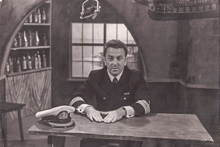 Mężczyzna w mundurze marynarskim siedzi za stołem. W tle sceneria portowej tawerny