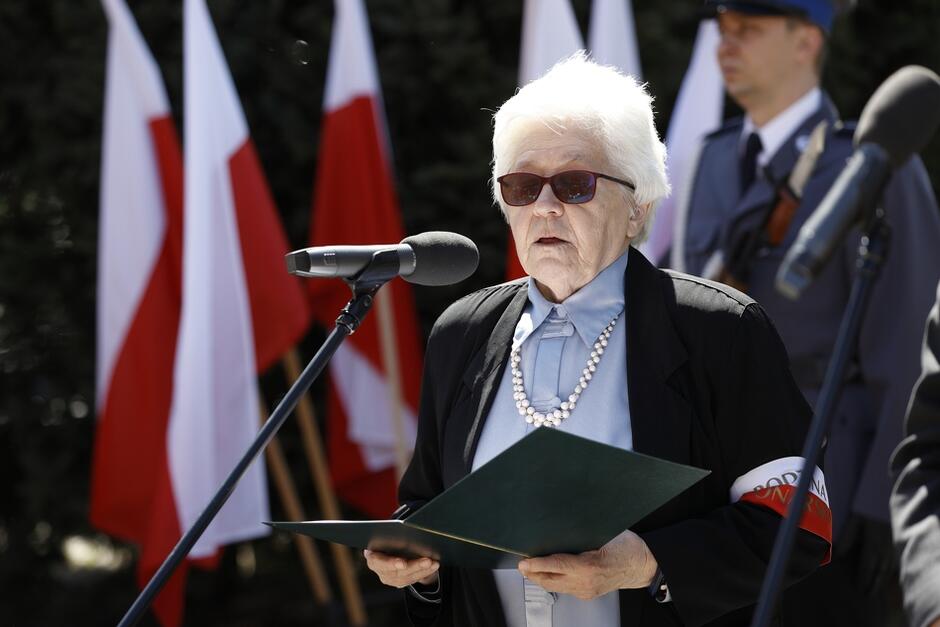 starsza kobieta, z siwymi włosami czyta przemówienie na tle pomnika