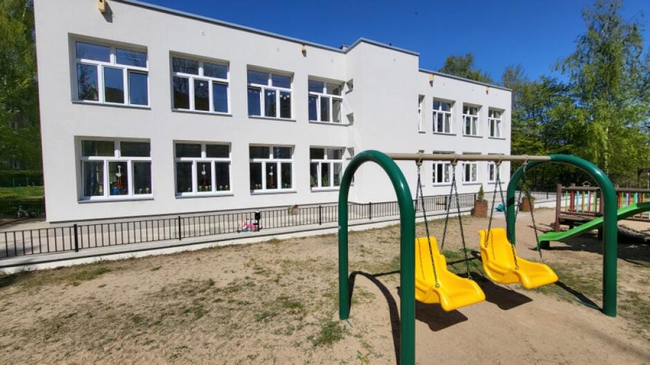 na zdjęciu na pierwszym planie zielono żółta huśtawka dla dzieci, w tle jasny dwukondygnacyjny budynek