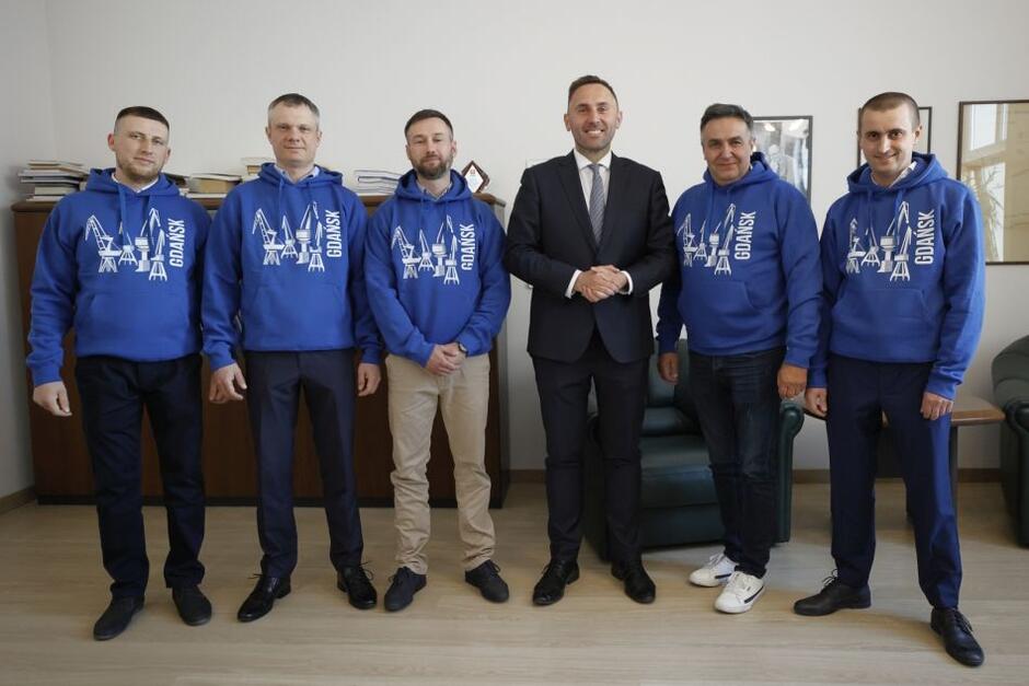 pięciu mężczyzna na w niebieskich bluzach z dźwigami, stoi obok mężczyzny w średnim wieku w garniturze