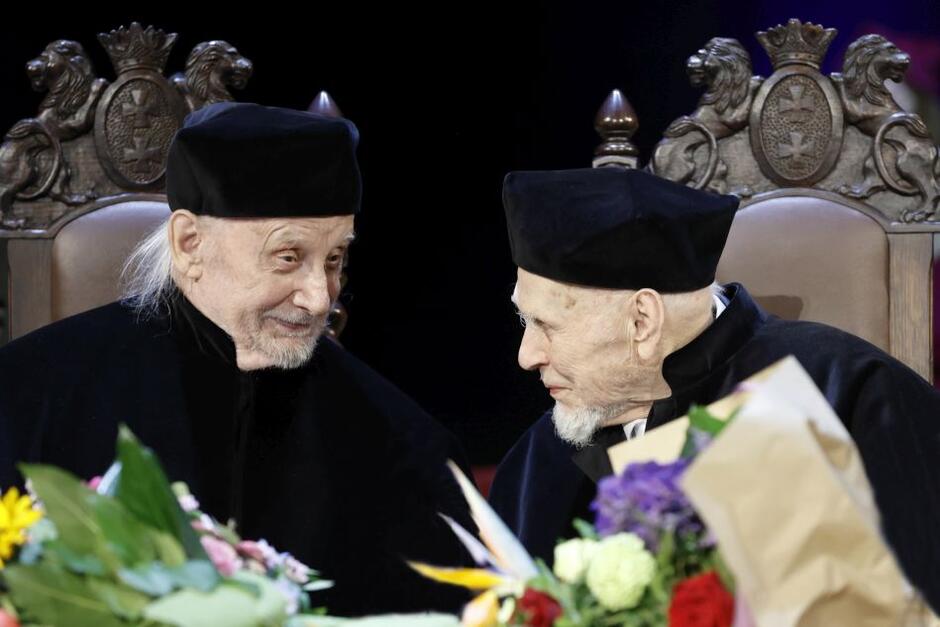 dwóch uśmiechniętych starszych mężczyzn w czarnych togach zwraca się do siebie