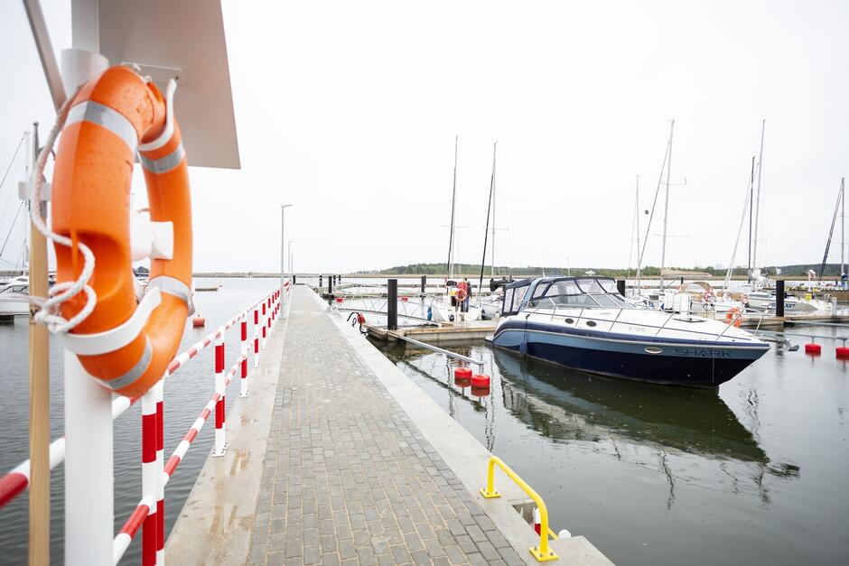 na zdjęciu fragment portu jachtowego, na pierwszym planie pomarańczowe koło ratunkowe po lewej, a po prawe zacumowana łódź