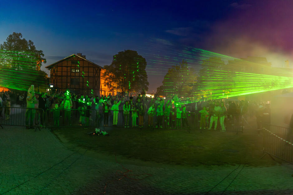 polana po zmroku, na niej ludzie, z prawej strony oświetla ich laserowa wiązka zielonego światła