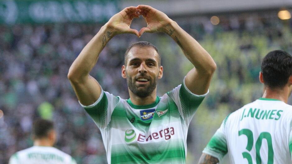 Uśmiechnięty mężczyzna w biało-zielonej koszulce. Dłonie trzyma nad głową w kształcie serca