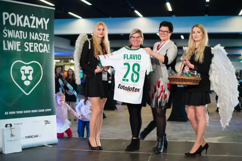 Cztery kobiety stoją obok siebie, trzymają koszulkę z nr 28 i napisem FLAVIO