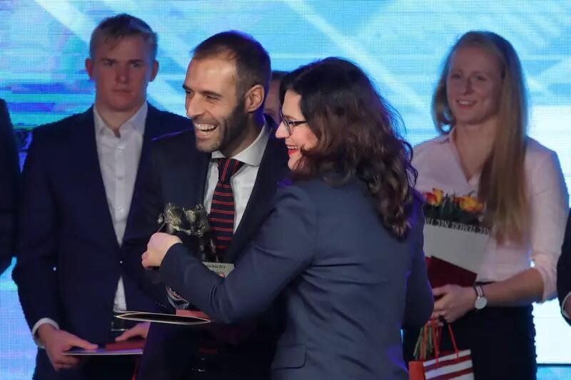 Uśmiechnięty mężczyzna w garniturze, obok uśmiechnięta kobieta w garsonce. Mężczyzna trzyma dyplom i statuetkę. W tle inne osoby