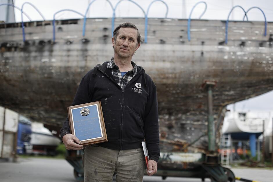 Mężczyzna stoi na tle jachtu, w ręce po lewej trzyma coś w rodzaju prostokątnego granatowego kafla w brązowej ramce 
