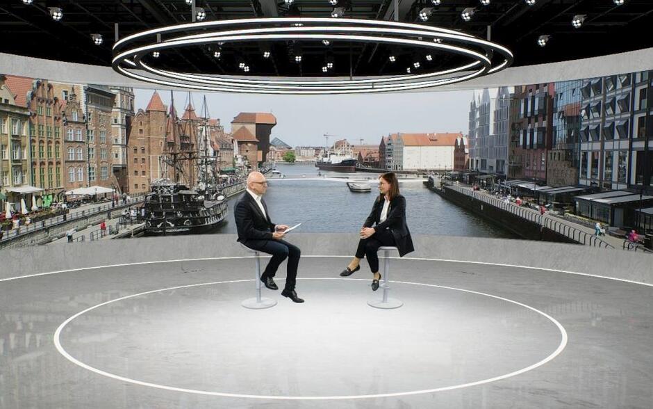 kobieta i mężczyzna w wirtualnym studiu, siedzą i rozmawiają, w tle widoki Gdańska