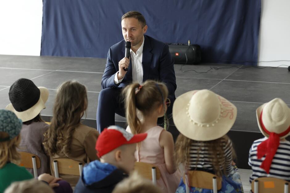 mężczyzna w garniturze mówi do mikrofonu do siedzących na widowni dzieci