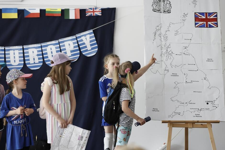dzieci w wakacyjnych strojach na scenie, jedna dziewczynka pokazuje palcem na rysunkową mapę Wielkiej Brytanii